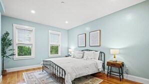 A bedroom with a bed, two bedside tables, a nightstand, a floor lamp, a potted plant, and two windows with blinds. - Modern Cozy 3BR-2BA Apt Near T (Boston)