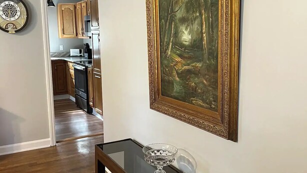 A hallway with a wooden floor, a glass-top table with a decorative bowl, and a framed painting of a forest scene. - Mountain Mama (Hendersonville)