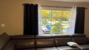 A living room with a brown leather sofa, a window with black curtains, and a view of a residential area with trees and parked cars. - Mountain Mama (Hendersonville)