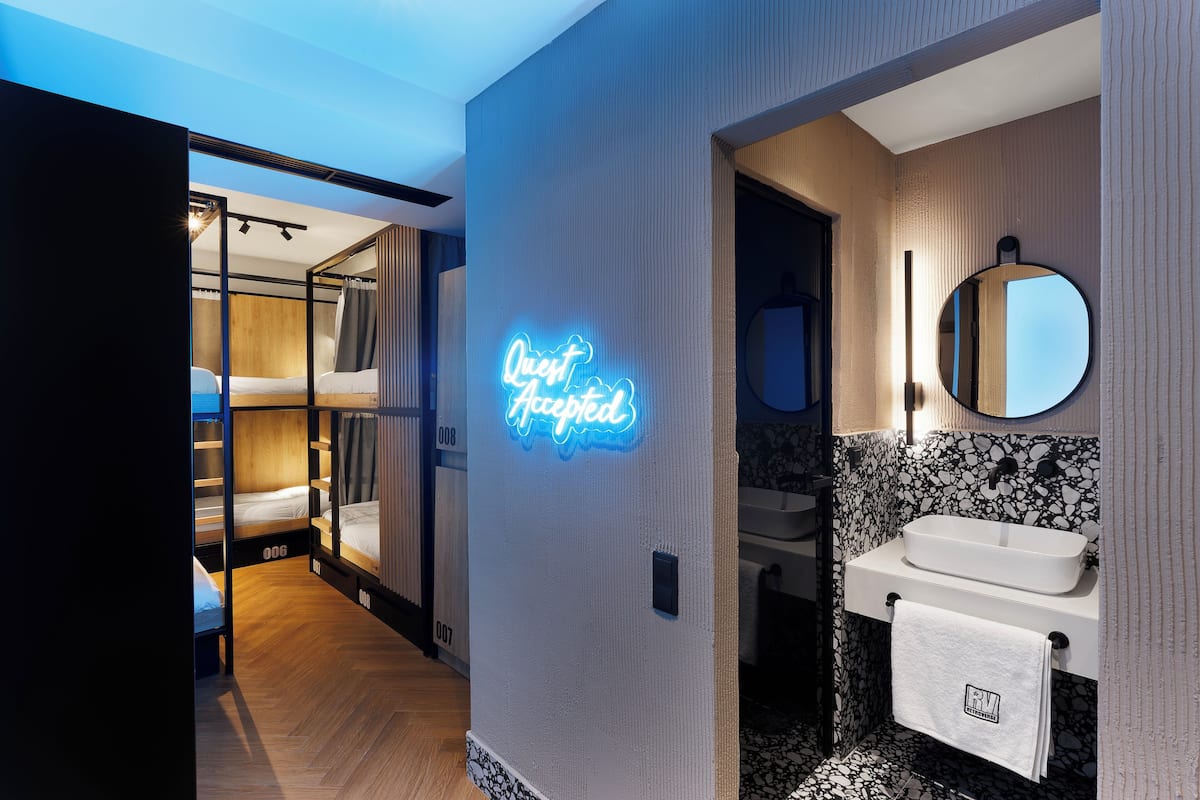 A modern hotel room with a neon sign, bunk beds, a bathroom with a sink and mirror, and a patterned backsplash.