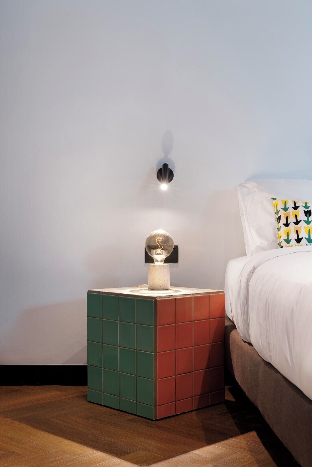 A neatly arranged bedroom with a bed, a nightstand with a lamp, and a wall-mounted light fixture.