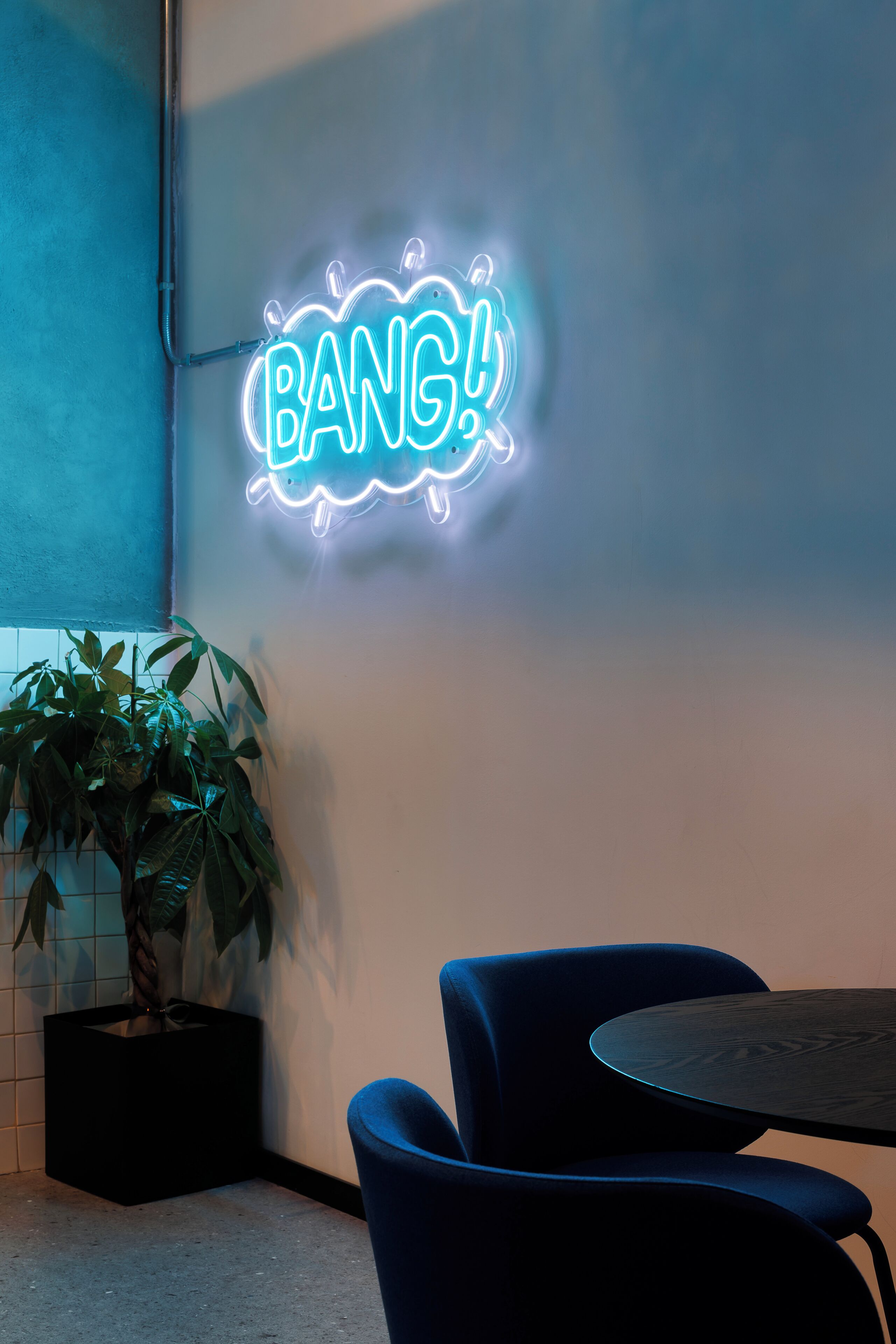 A neon sign with the word "BANG!" on a wall, a potted plant, and two blue chairs around a small table.