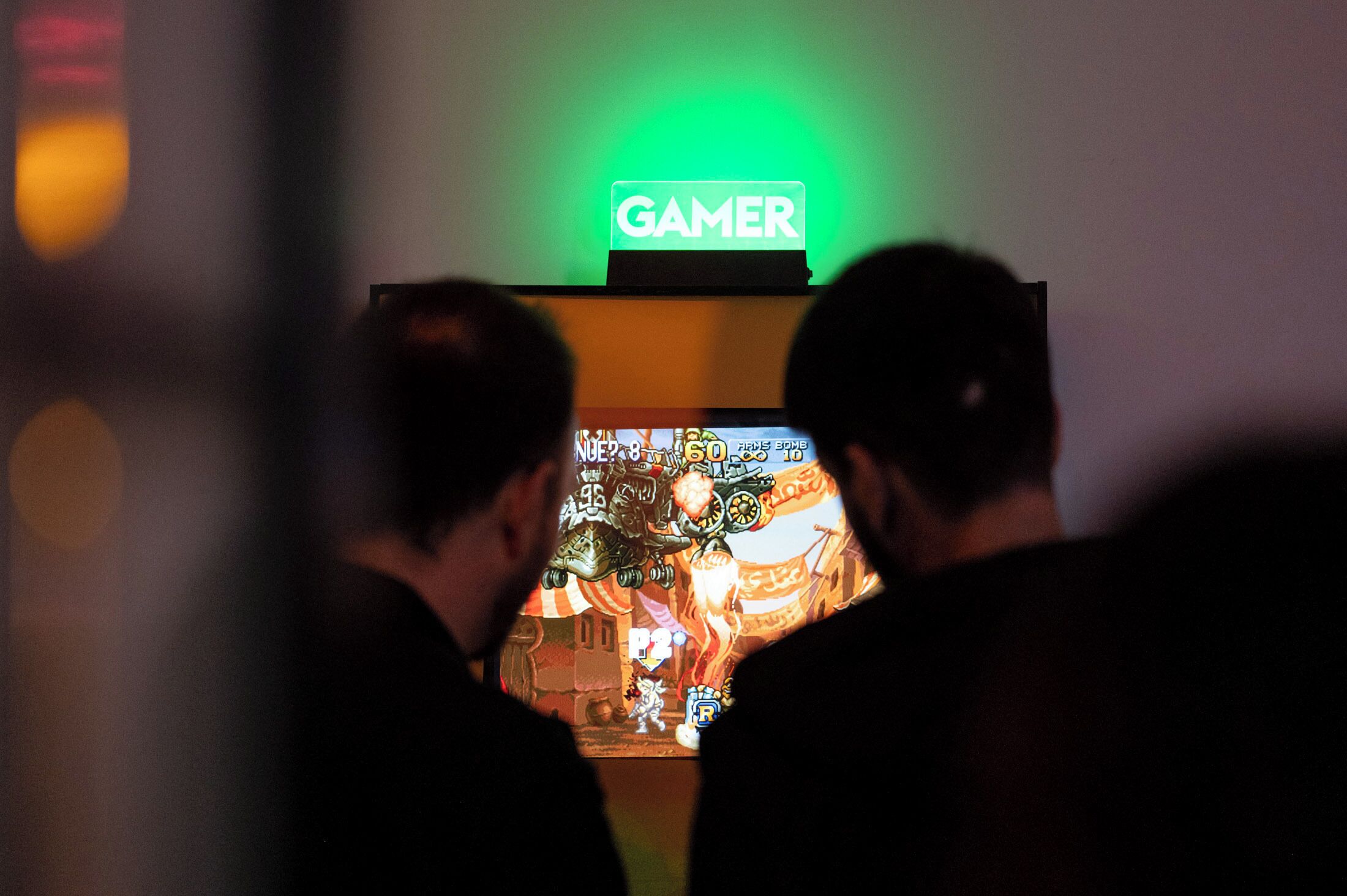 Deux personnes jouant à un jeu vidéo, avec un panneau lumineux affichant le mot « GAMER » au-dessus d’elles.