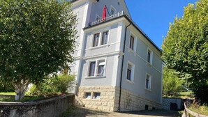 Ein zweistöckiges Gebäude mit rustikalem Steinunterbau und weißen Obergeschossen, einer roten Markise und einem gepflasterten Auffahrt.