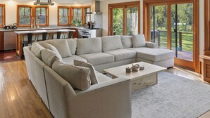 A modern living room with a large L-shaped sectional sofa, a coffee table, and a kitchen area with wooden cabinets and a large window.