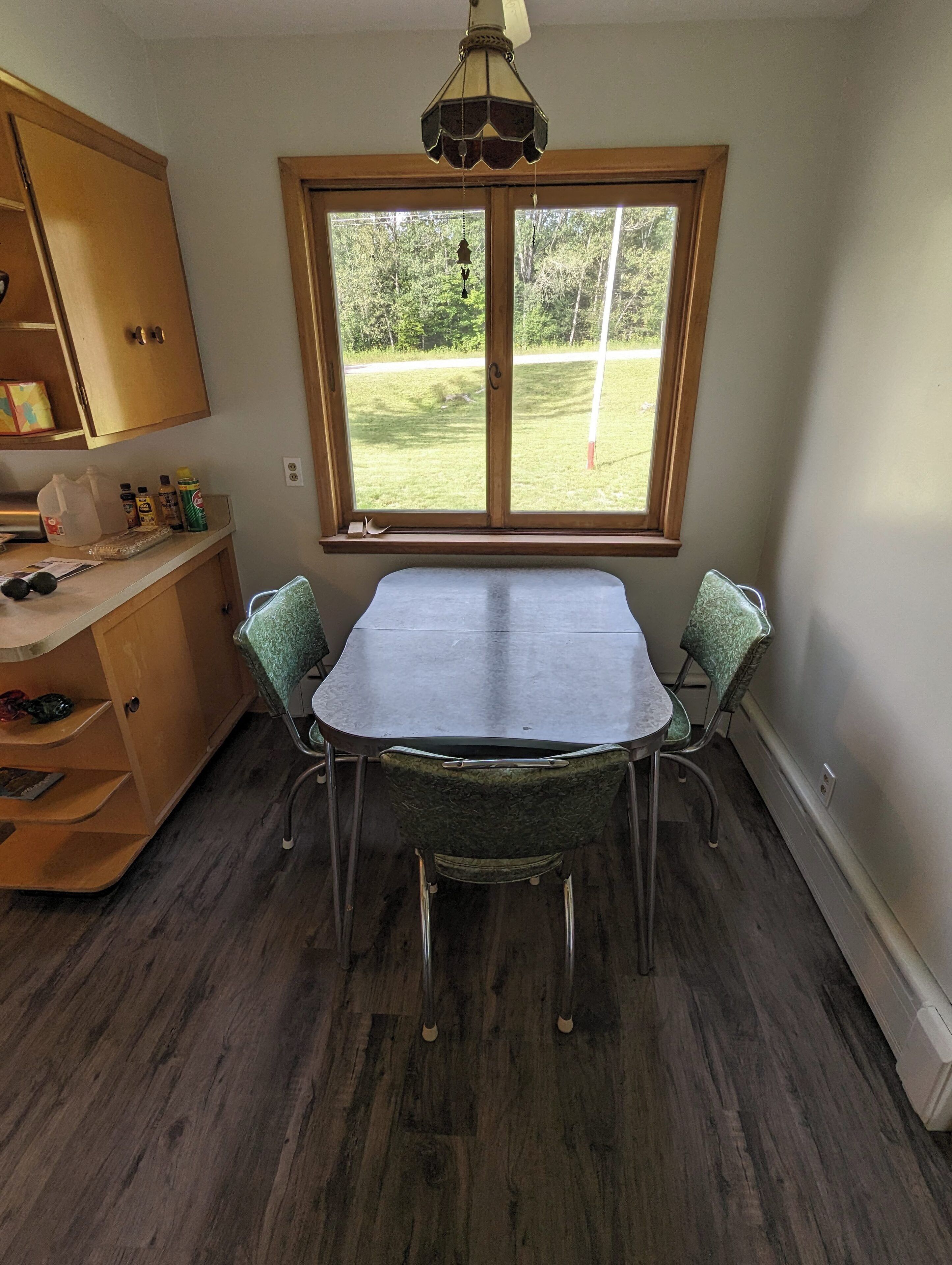 Un comedor con una mesa y sillas, un ventanal con vistas a una zona verde, armarios de madera y una lámpara en el techo.
