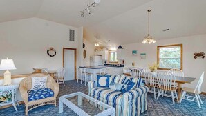 A spacious living area with a sofa, chairs, a dining table, and a kitchen in the background. - Oceanside 5BR pet-friendly Corolla home with private pool & hot tub! (Corolla)