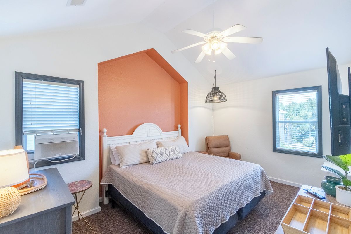 A bedroom with a bed, a ceiling fan, a bedside table with a lamp, a chair, and a window.