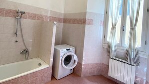 Casa de banho com banheira, máquina de lavar roupa, radiador e janela com cortinas.