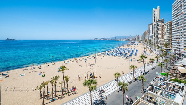 Una playa con hileras de sombrillas azules y palmeras, un paseo marítimo con gente caminando, y edificios altos al fondo.