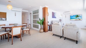Una sala de estar moderna con comedor, un ventanal grande y un televisor que muestra una escena de playa.