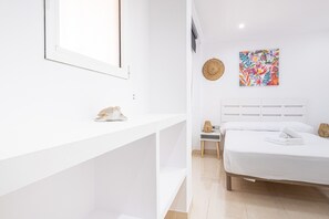 Un dormitorio moderno con cama blanca, una mesita de noche, una pequeña cómoda, una obra de arte en la pared y un estante blanco con un objeto decorativo.
