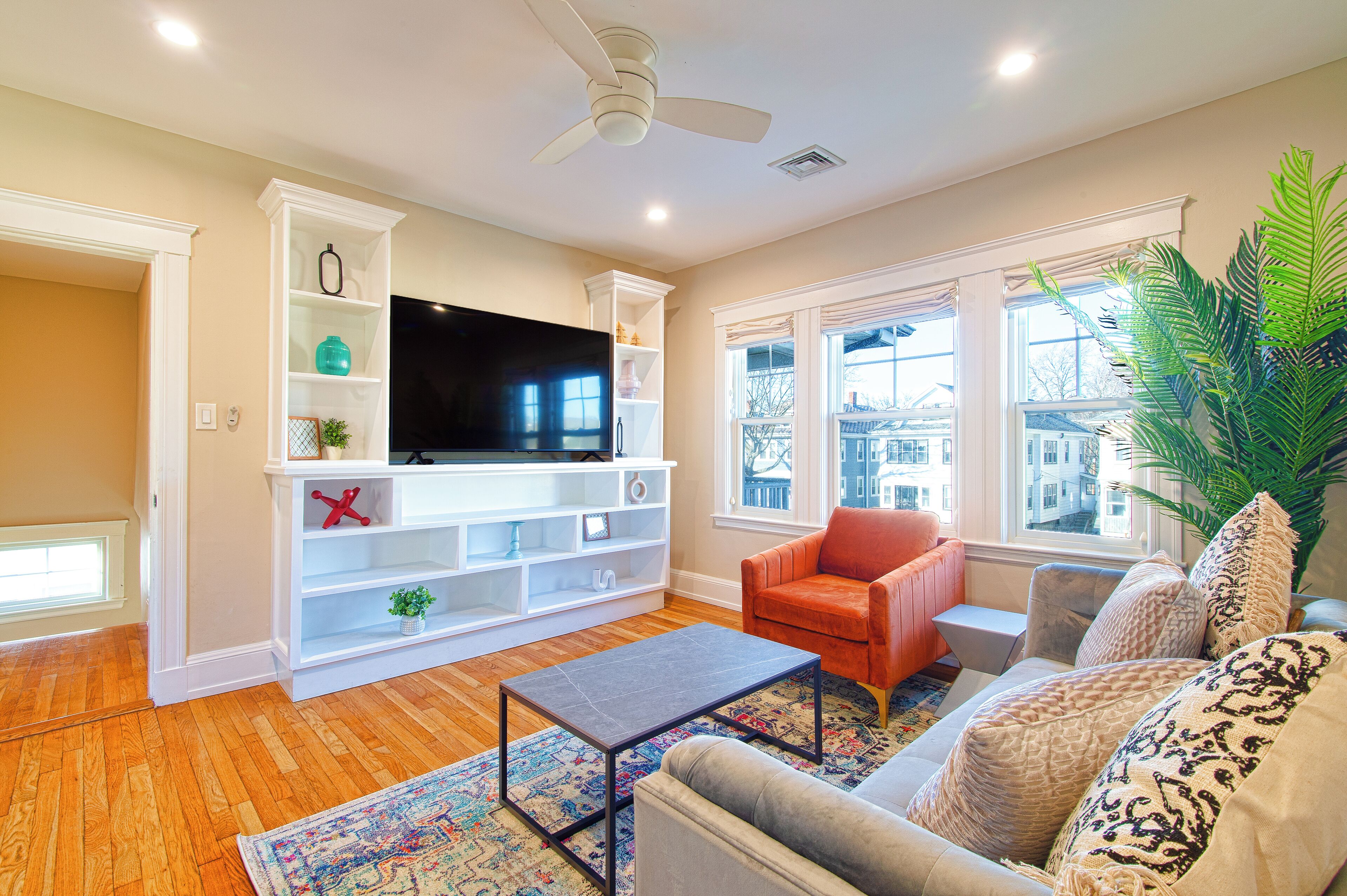 A living room with a flat-screen TV, white shelving, a red armchair, a gray sofa, and a large plant.