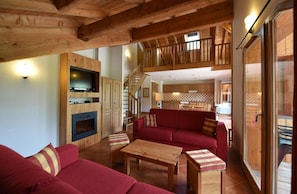 A spacious living area with a red sofa, wooden coffee table, and a fireplace. A kitchen with wooden cabinets and a dining area is visible in the background.