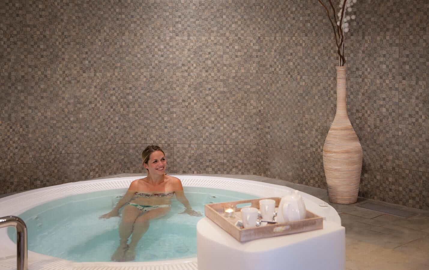 A woman in a hot tub with a tray of amenities.