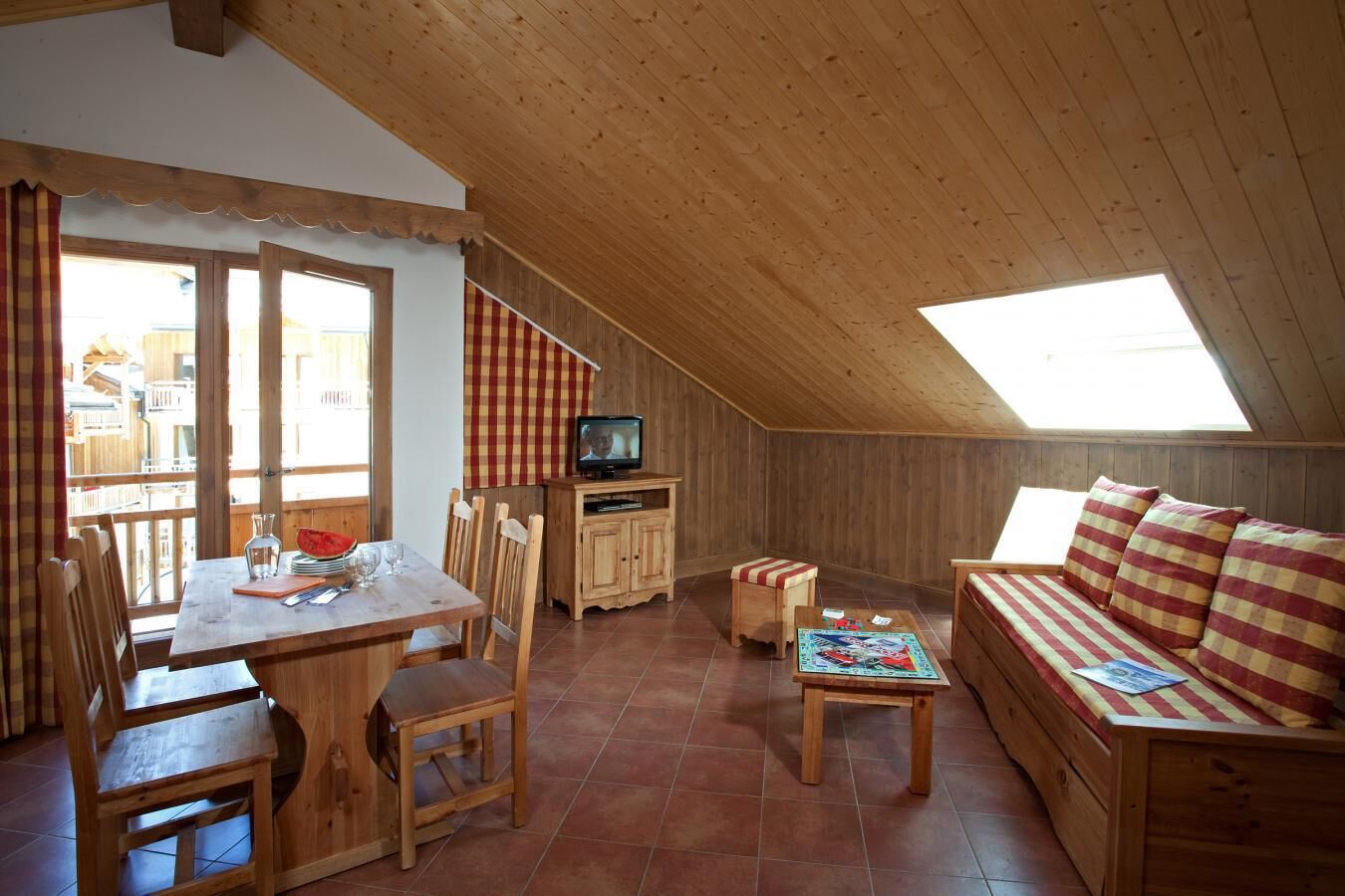 A cozy attic room with a wooden ceiling, a dining table with chairs, a sofa, and a television.