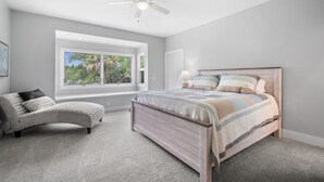 Ein Schlafzimmer mit großem Fenster, Deckenventilator, Bett mit gestreifter Decke und einem Sessel.