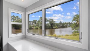 Ein Zimmer mit großen Fenstern, die auf einen See und Bäume blicken, eine weiße Bank und eine deckeneinliegende Lampe.