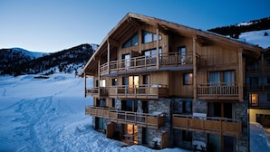 Un chalet en bois à plusieurs étages, doté de balcons, agrémenté de pierres apparentes, et avec une piste de ski en arrière-plan.