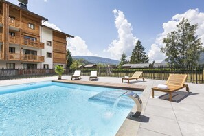 A swimming pool with lounge chairs, a wooden building with balconies, and a mountainous backdrop. - Wifi, swimming pool, terrace, balcony, tv, ski locker, 34-44m², Samoëns (Samoëns)