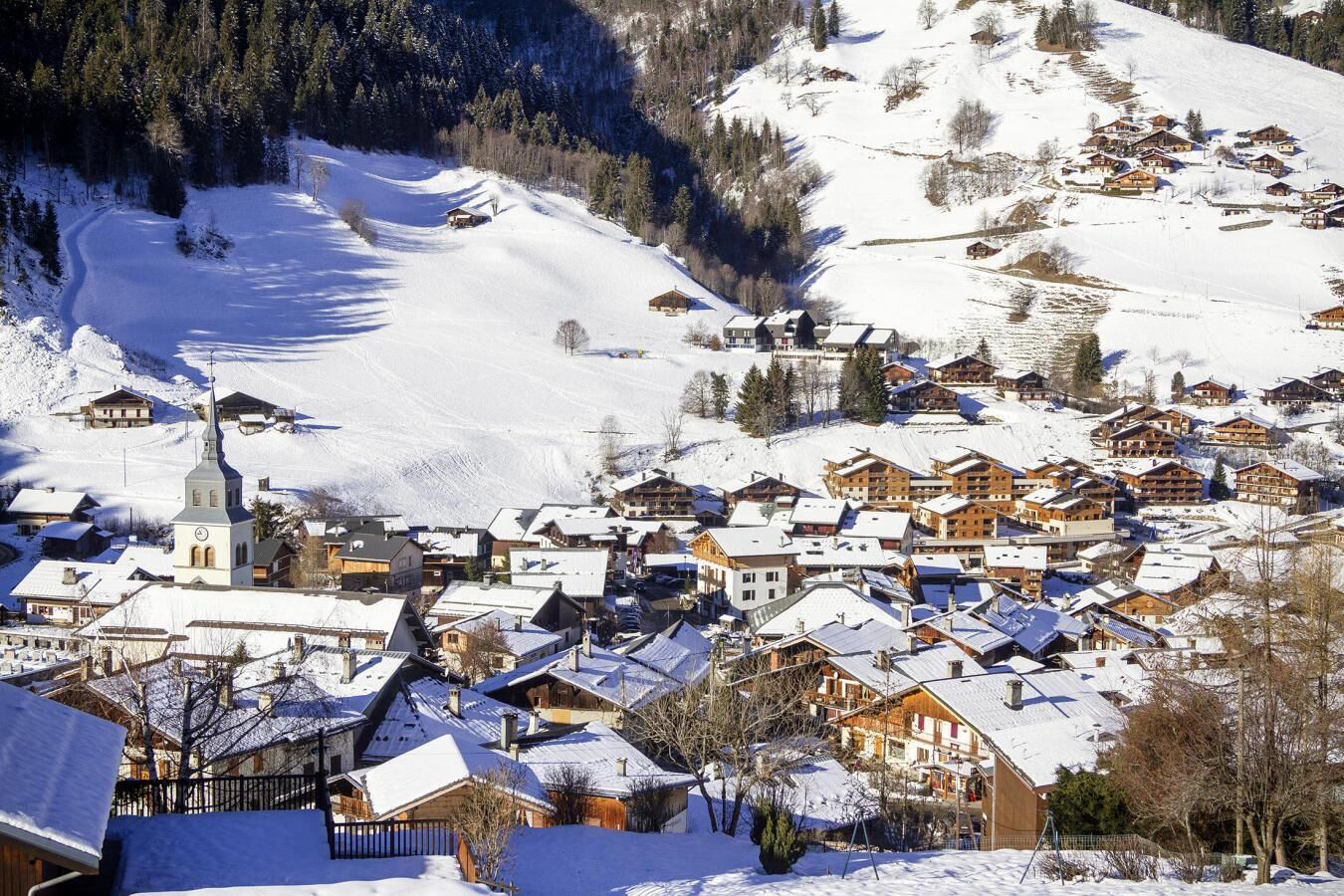 A snow-covered village with traditional wooden houses, a church, and a ski slope in the background.
