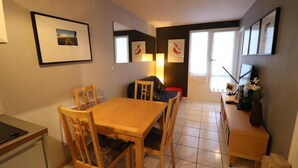 Un rincón comedor compacto con una mesa y sillas de madera, una kitchenette pequeña y un televisor sobre un mueble.
