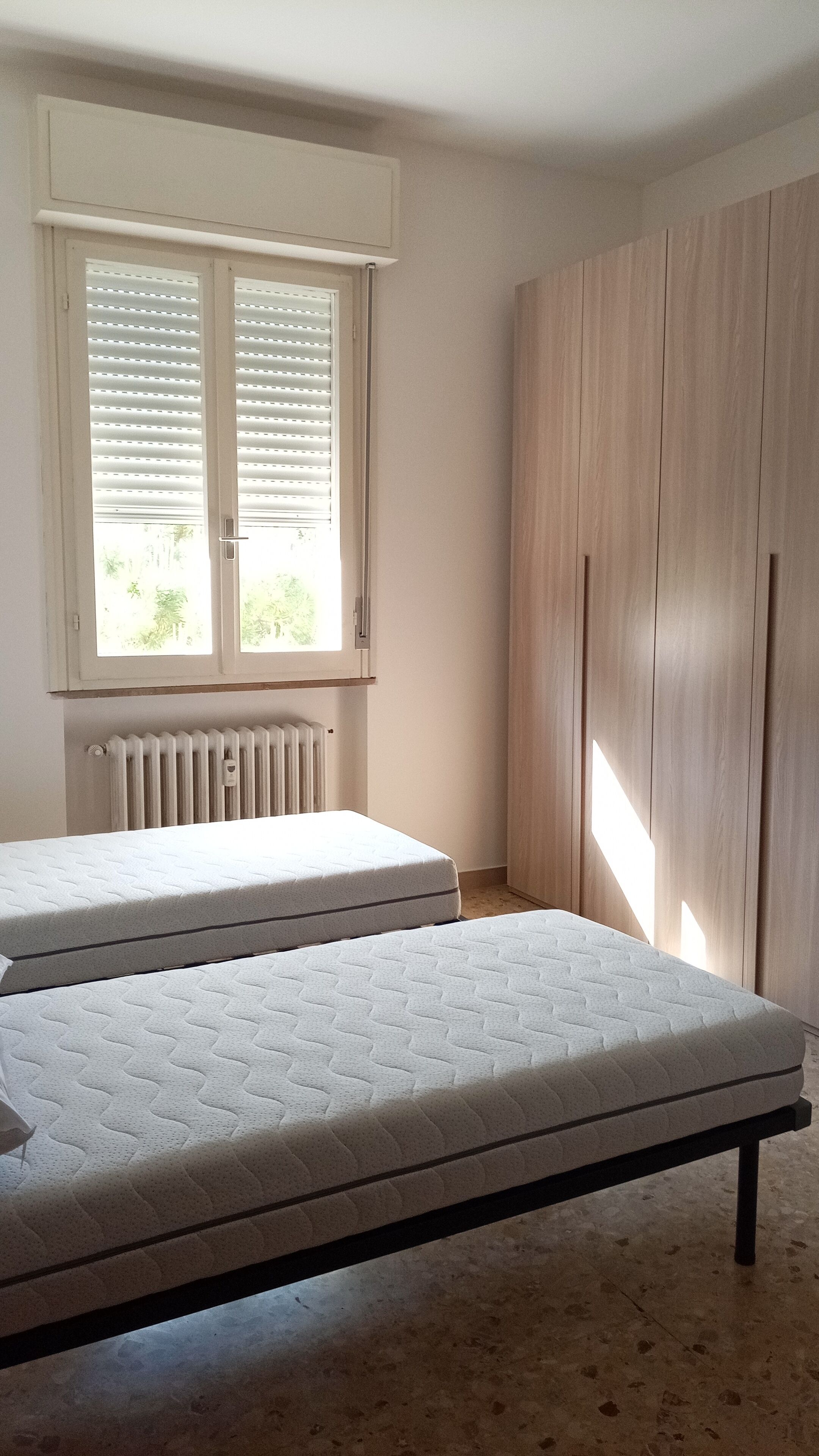 Quarto com duas camas de solteiro, janela com persianas, radiador e guarda-roupa de madeira.