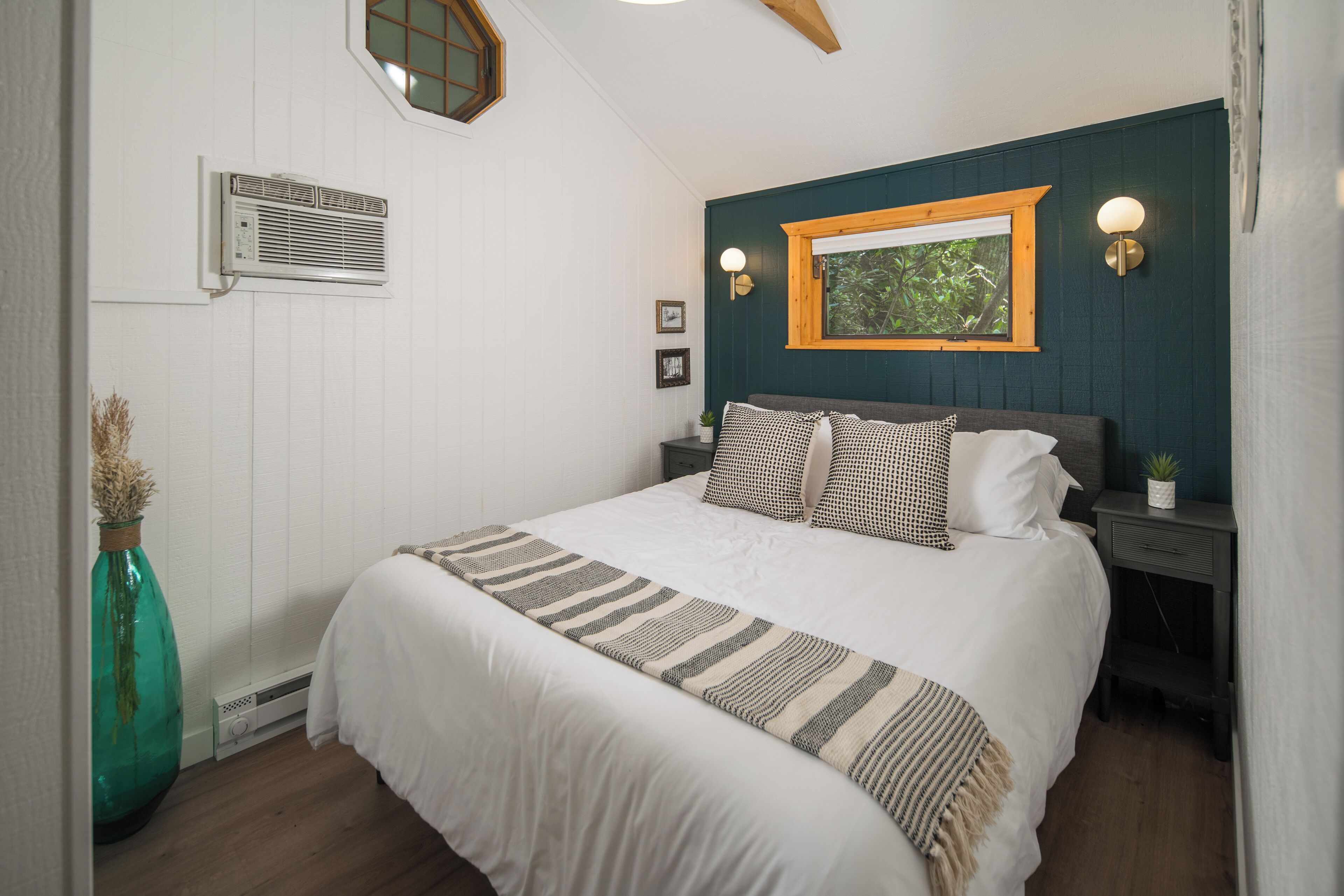 A bedroom with a large bed, a window with a view, a wall-mounted air conditioner, and a bedside table with a lamp.