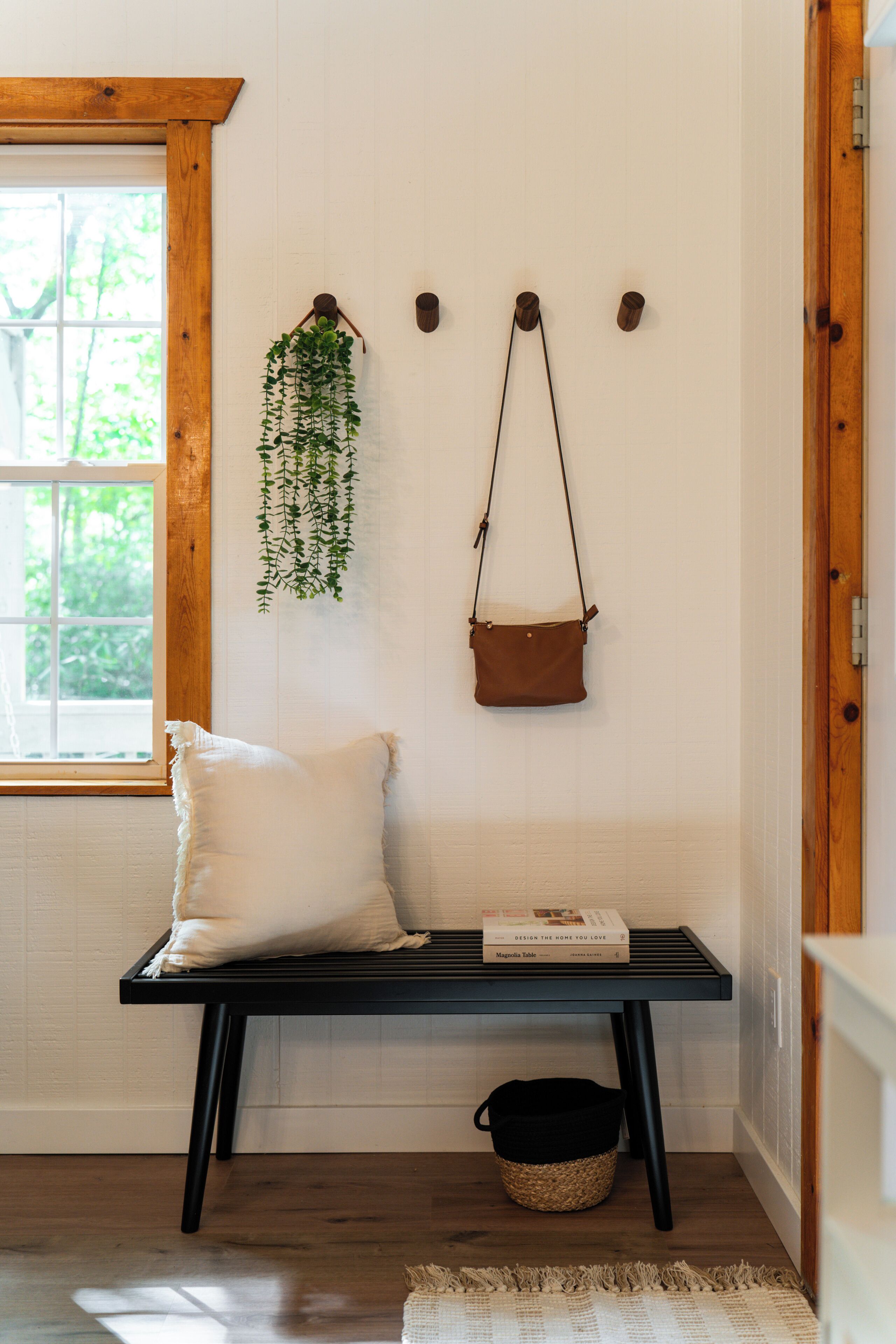 A room with a wooden-framed window, a white wall, a black bench with a white pillow, a hanging plant, and a brown bag on hooks.