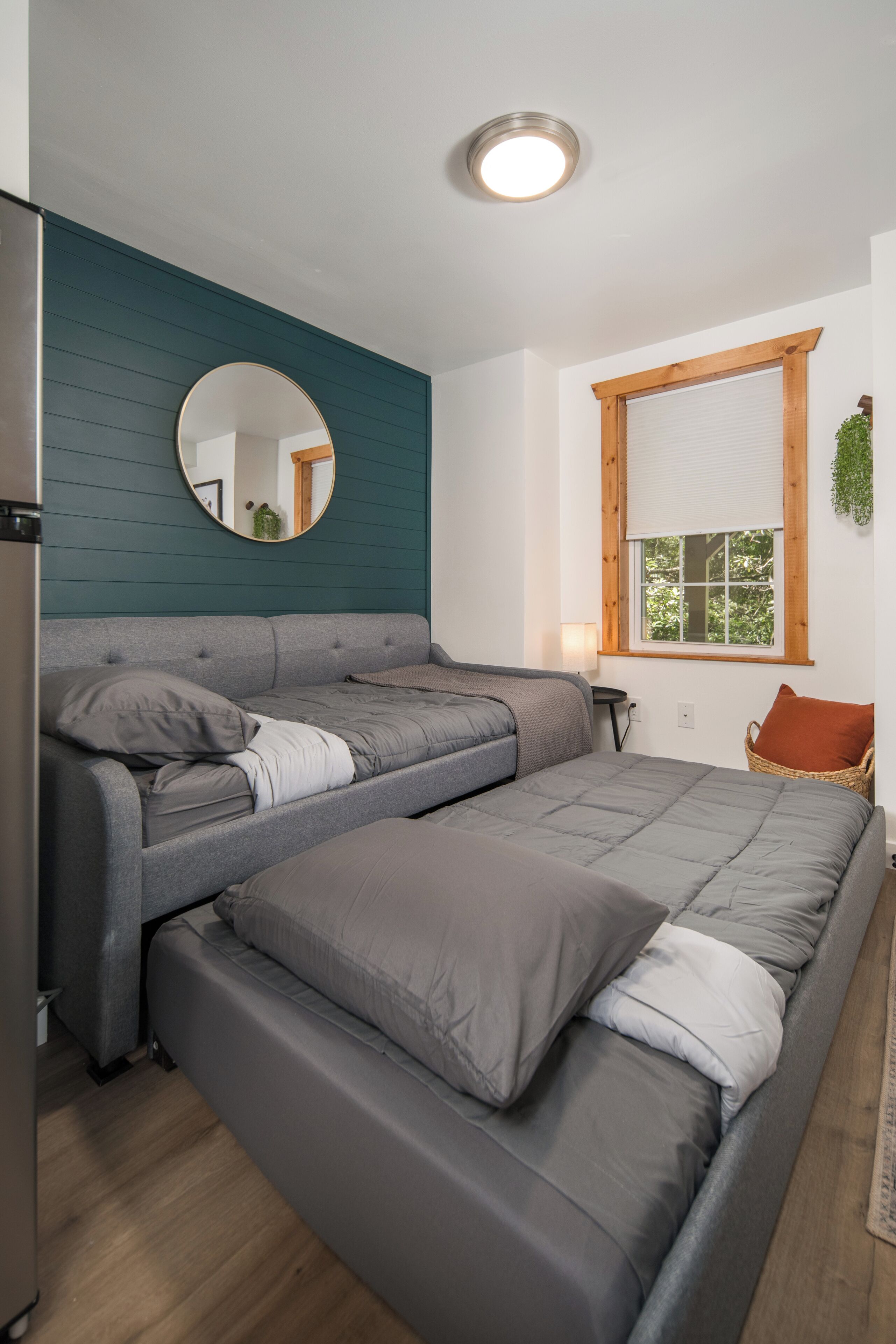 A room with a sofa bed, a round mirror, a window with blinds, and a small table with a lamp.