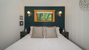 Een net opgemaakt bed met twee kussens, een nachtkastje met een lamp en een raam met uitzicht op bomen.