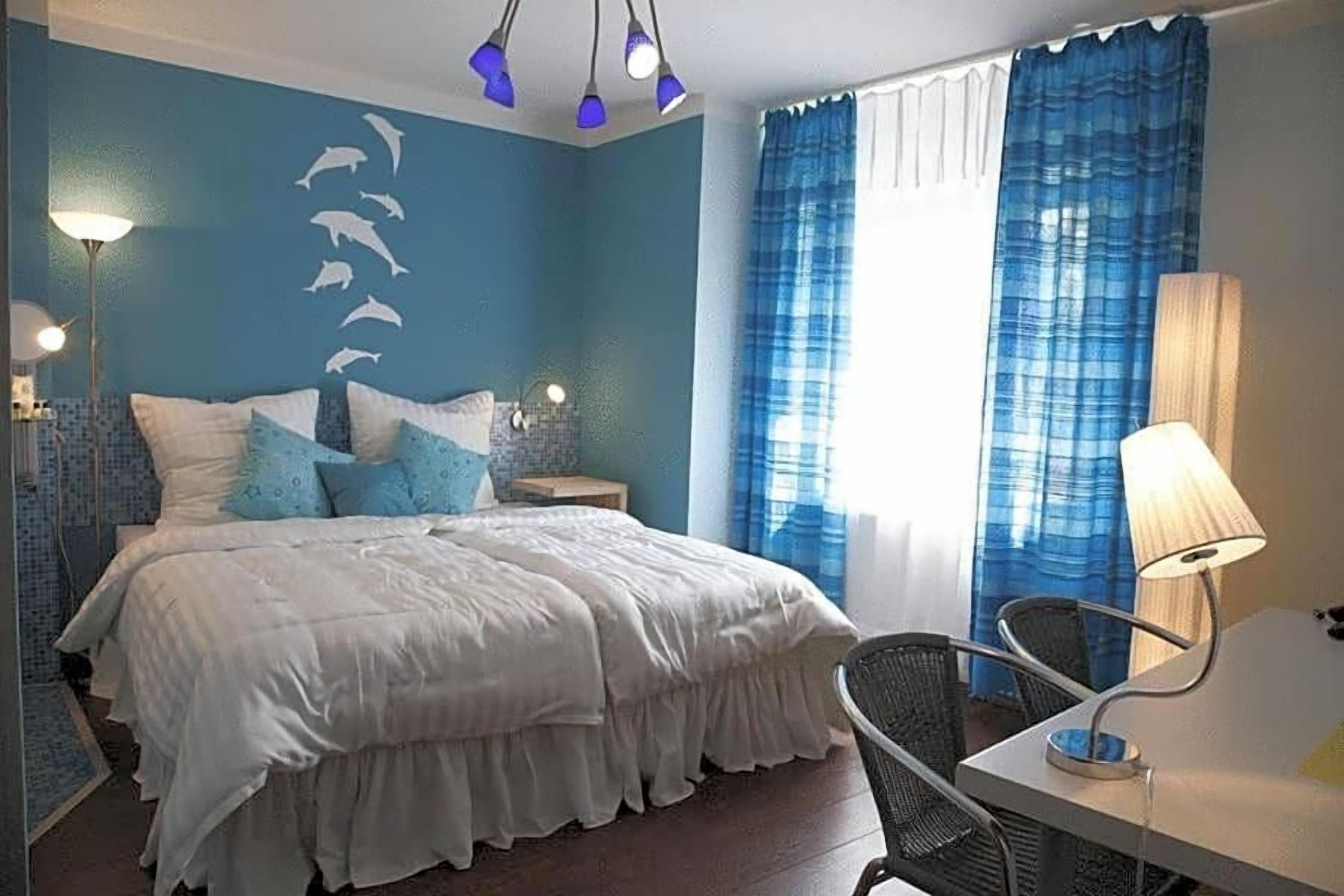 A bedroom with a bed, a chair, a nightstand, a lamp, and a wall with dolphin silhouettes.