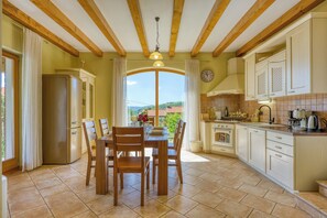 A kitchen with wooden beams, a dining table with chairs, a refrigerator, and a view through a window.