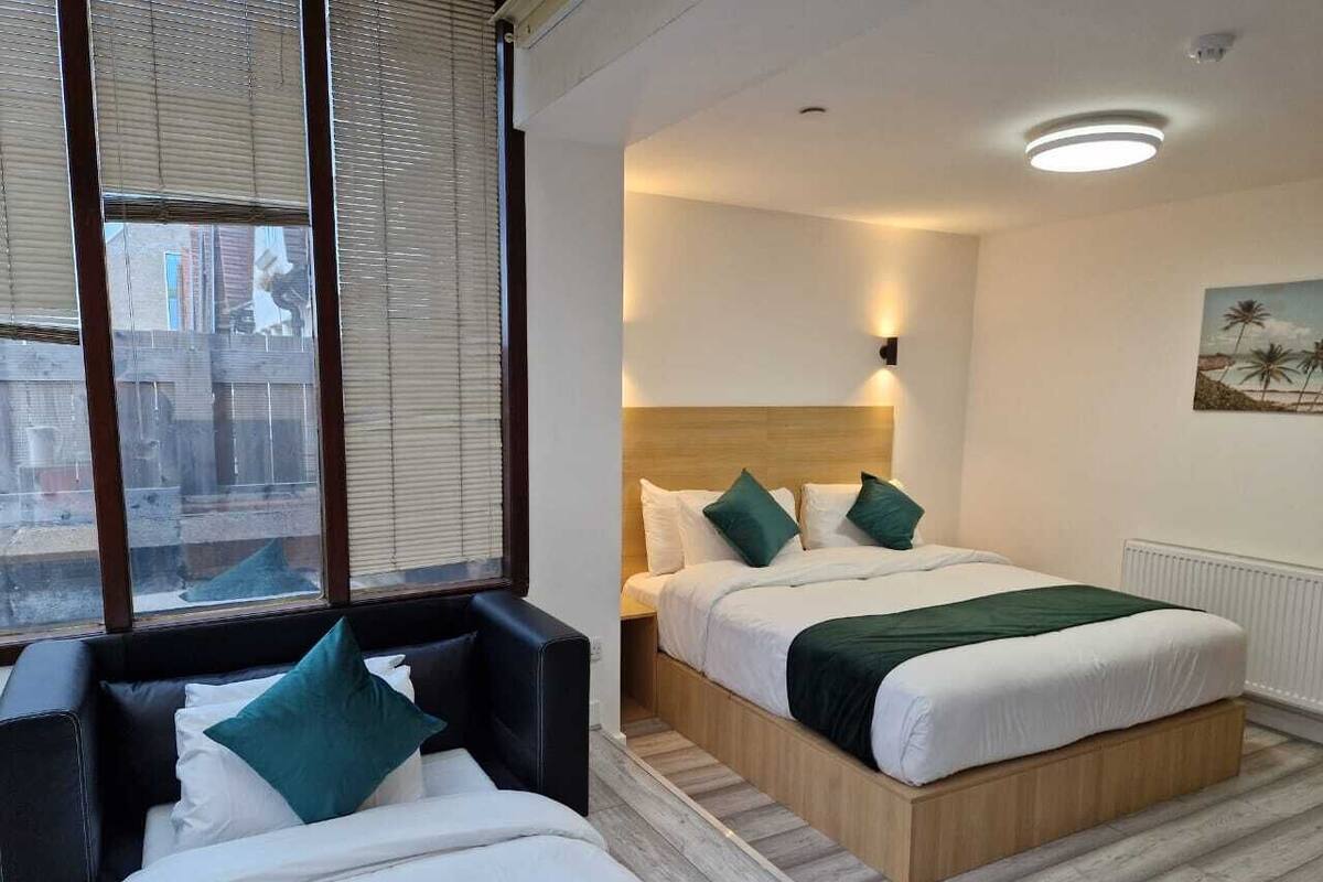 A hotel room with a bed, a sofa, a window with blinds, and a painting on the wall.
