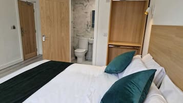 A bedroom with a bed, two pillows, a wooden wardrobe, and a bathroom visible through an open door.