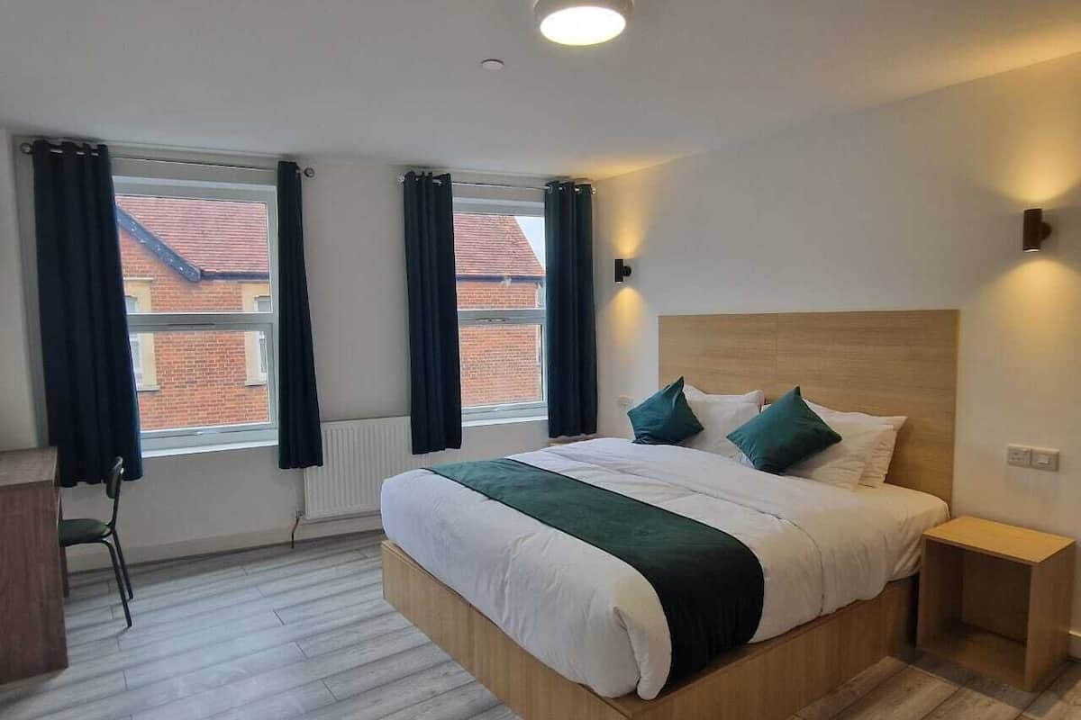 A modern bedroom with a large bed, a wooden headboard, a bedside table, and a view of a brick building through the window.