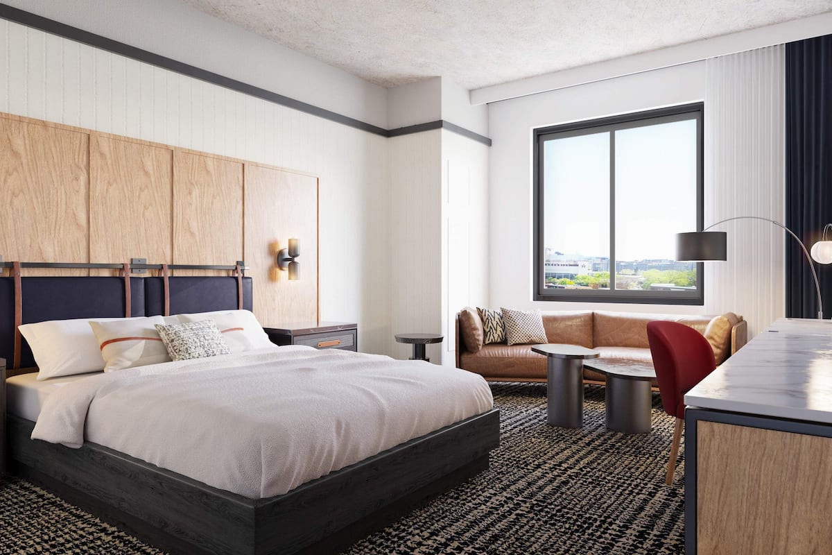 Ein modernes Hotelzimmer mit einem großen Bett, einer Couch, einem kleinen Tisch und einem Schreibtisch. Es gibt ein Fenster mit Blick auf die Stadt, eine Wandleuchte und ein gerahmtes Bild an der Wand.
