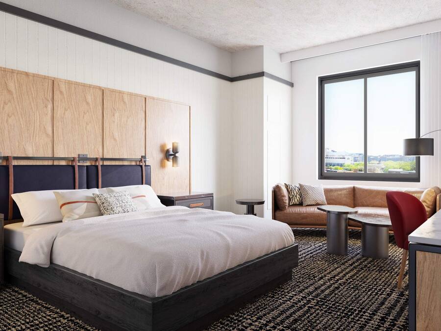 Una habitación de hotel moderna con una cama grande, un sofá, una mesita y un escritorio. Cuenta con una ventana con vistas a la ciudad, una lámpara de pared y un cuadro en la pared.