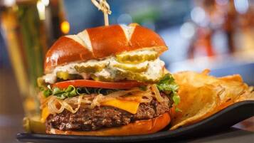 Hamburguesa con pepinillos, queso y cebolla, acompañada de papas fritas y una bebida al fondo.