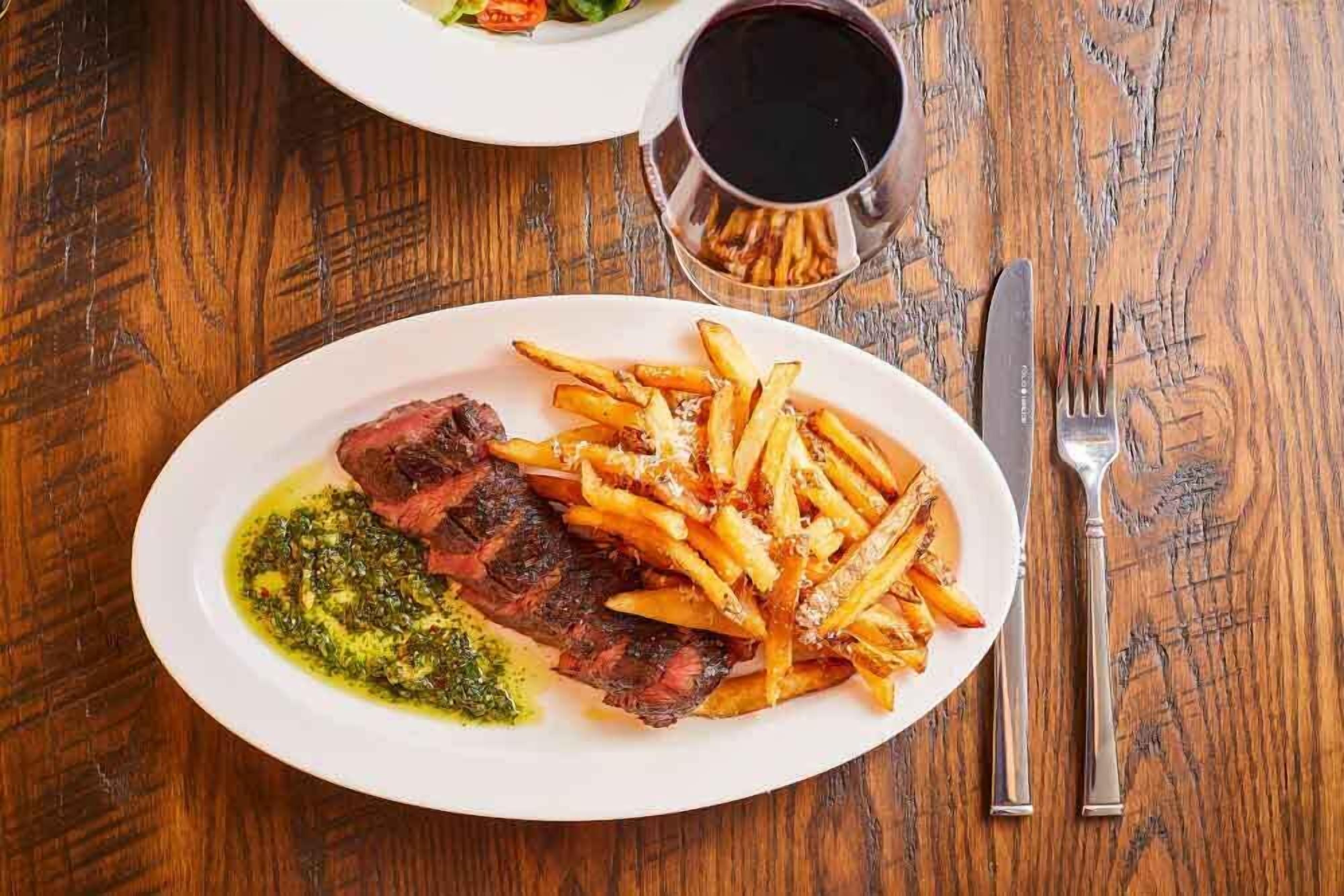 Un plato de carne con papas fritas y salsa verde, una copa de vino tinto y cubiertos sobre una mesa de madera.