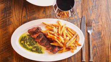 Un plato de carne con papas fritas y salsa verde, una copa de vino tinto y cubiertos sobre una mesa de madera.