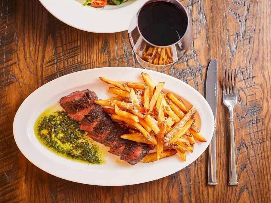A plate of steak with fries and a green sauce, a glass of red wine, and cutlery on a wooden table.