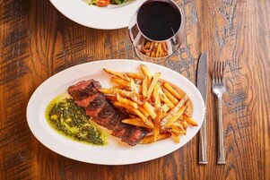 Un plato de carne con papas fritas y salsa verde, una copa de vino tinto y cubiertos sobre una mesa de madera.