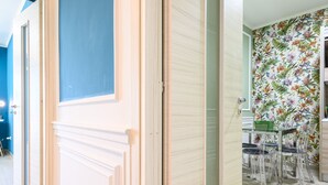 A hallway with a blue door, a wooden door, and a dining area with a floral wallpaper and clear chairs. - Via Nuova Marina 121 - Appartamento 1 (Naples)