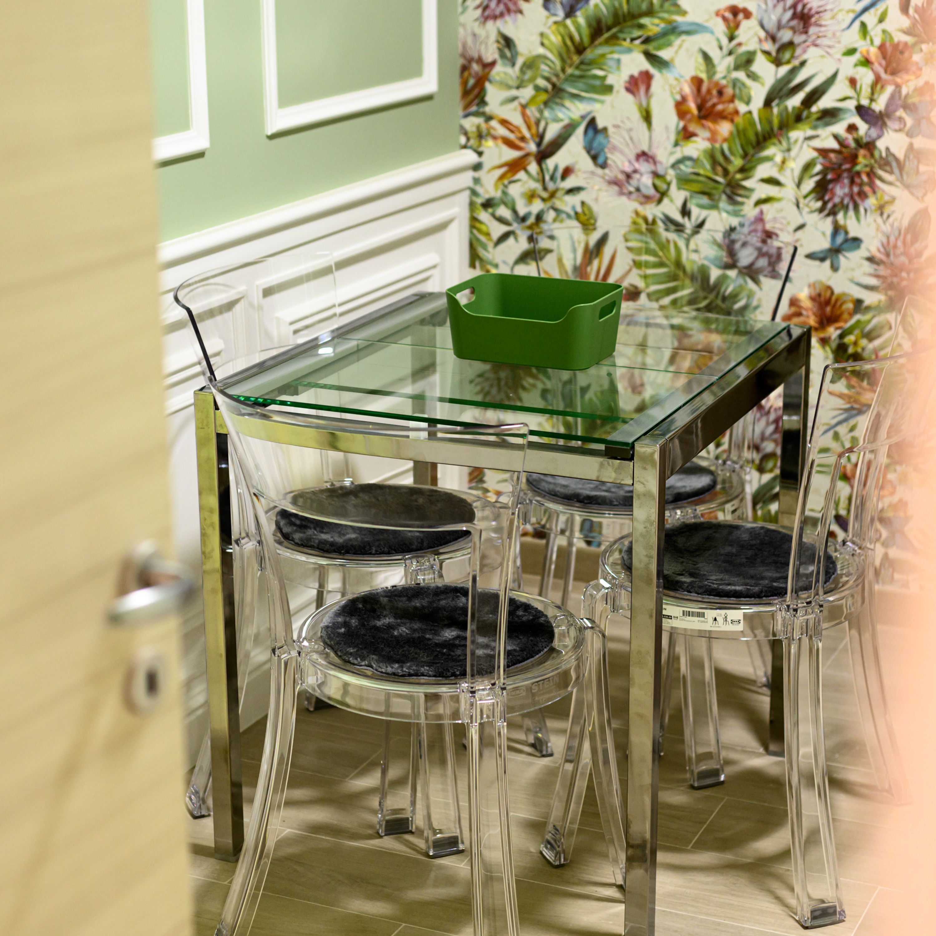 A clear glass dining table with four chairs, a green tray, and a floral wallpaper background.