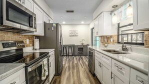 Una cocina moderna con armarios blancos, un refrigerador de acero inoxidable, un microondas y una estufa. El piso es de madera y hay una ventana con vistas al exterior.