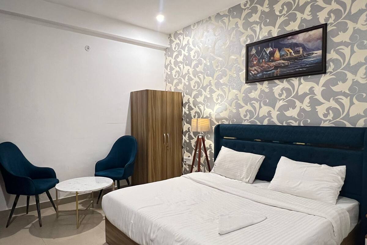 A hotel room with a bed, two chairs, a small table, a wardrobe, and a painting on the wall.