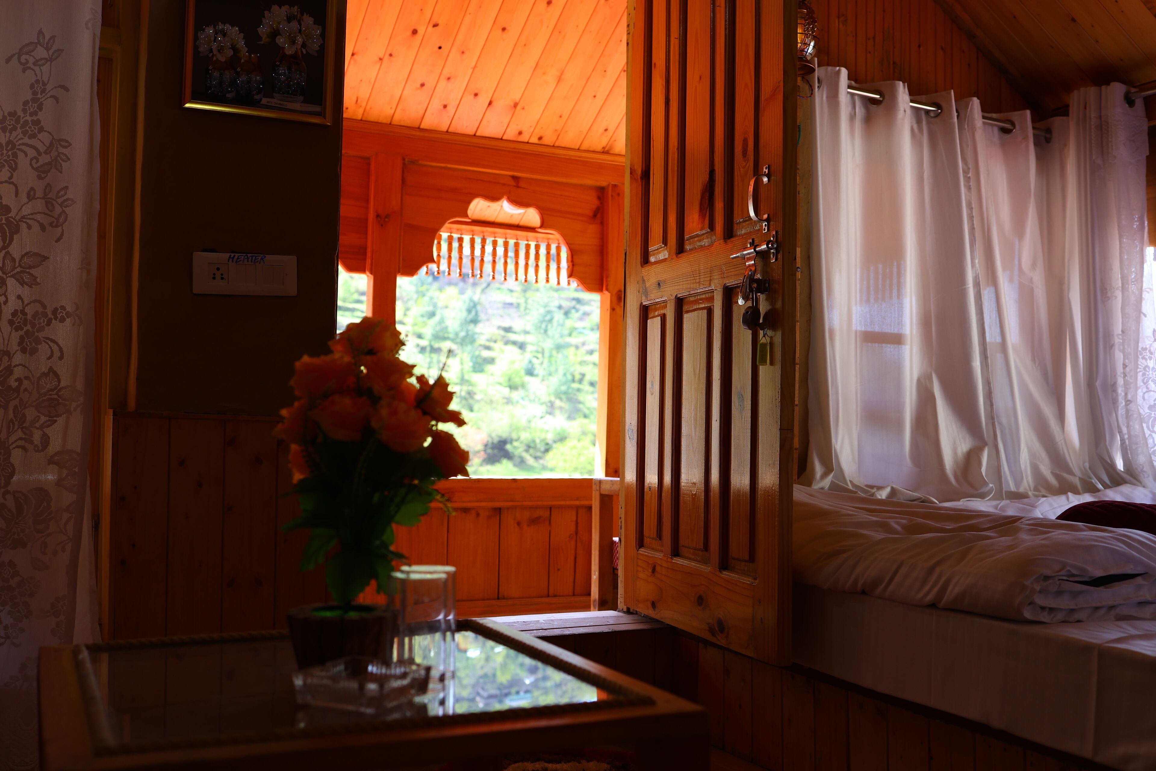 A wooden cabin room with a bed, a table with a vase of flowers, and a window with curtains.