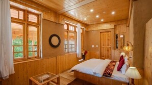 A wooden cabin bedroom with a bed, bedside table, and a window overlooking greenery. - Tirthan Village Resort (Banjar)