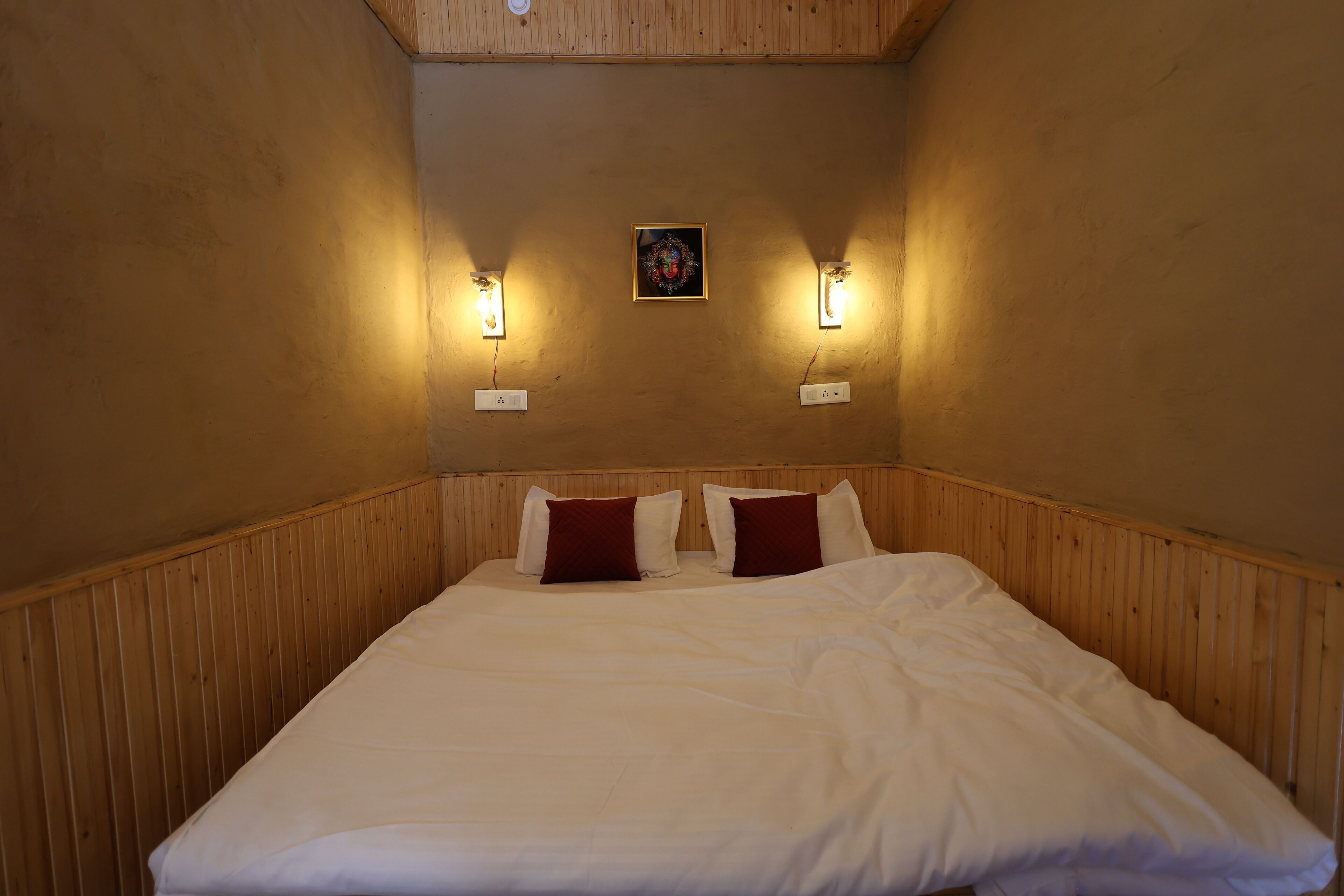 A single bed with white linens and two red pillows, flanked by two wall-mounted light fixtures, in a room with wooden paneling and a small framed artwork on the wall.